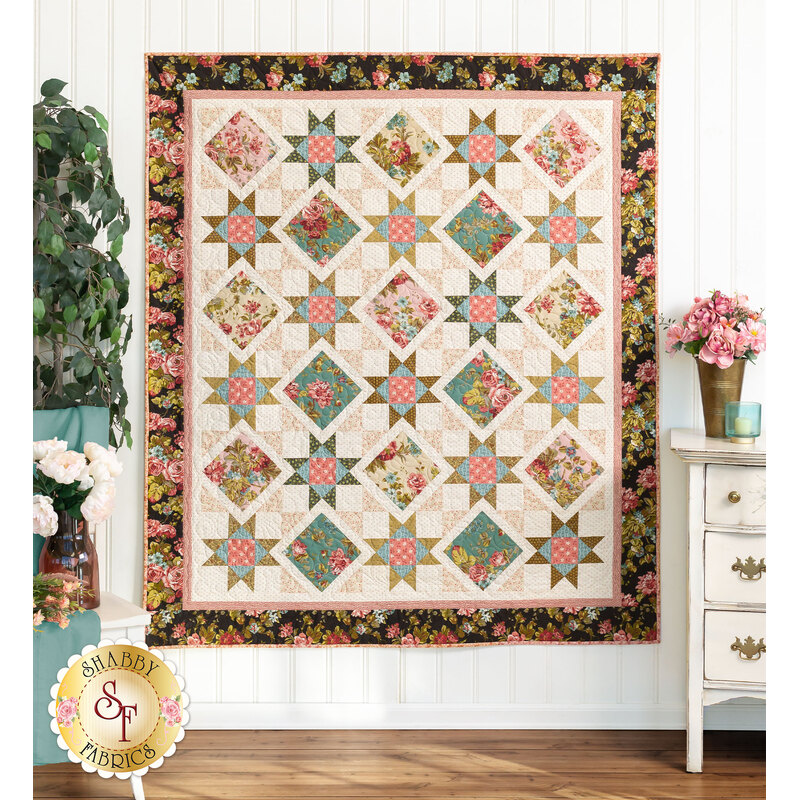 Photo of the finished quilt featuring black, teal, cream, and pink floral fabrics hanging on a white paneled wall with a houseplant, bouquets of flowers and a white antique dresser on the right.