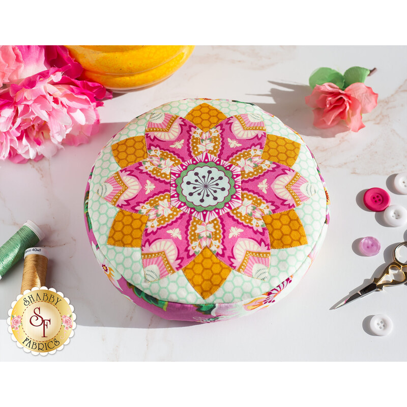 Image of a round yellow and pink pincushion quilted in an intricate flower shape on a white countertop with green and gold thread to the left of it, gold scissors and pink and white buttons to the right, and a pink flower in the upper left and right corners