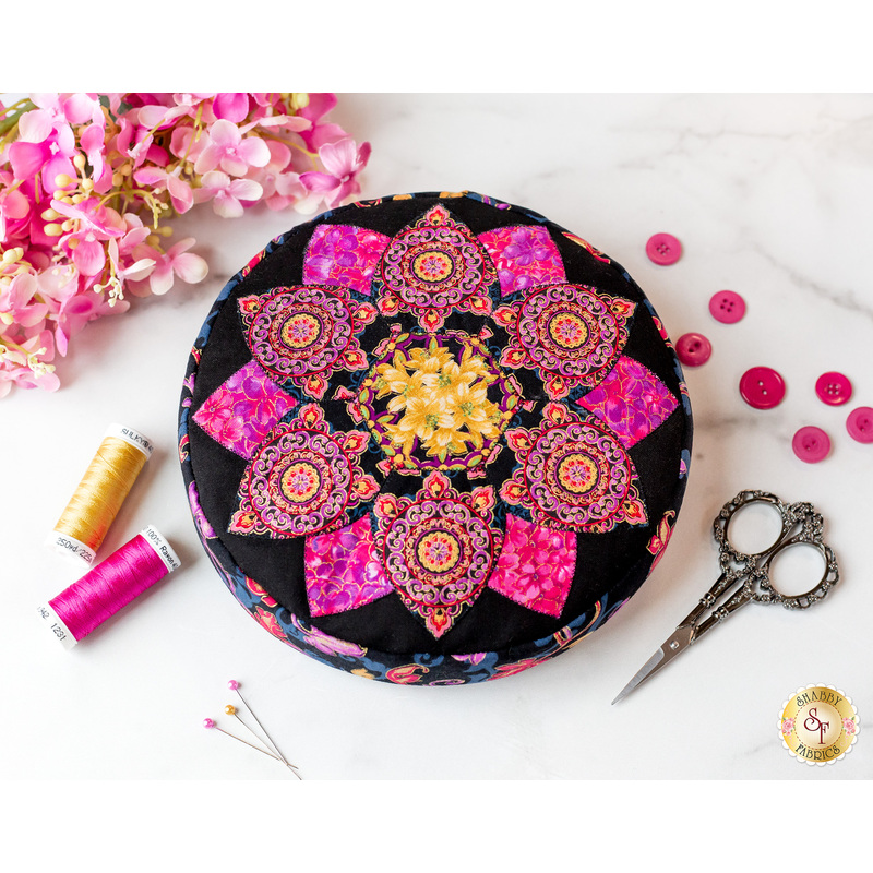 A round pink, yellow and black pincushion quilted in an intricate flower shape on a white marble countertop with pink and yellow themed notions scattered nearby and pink flowers in the top left corner