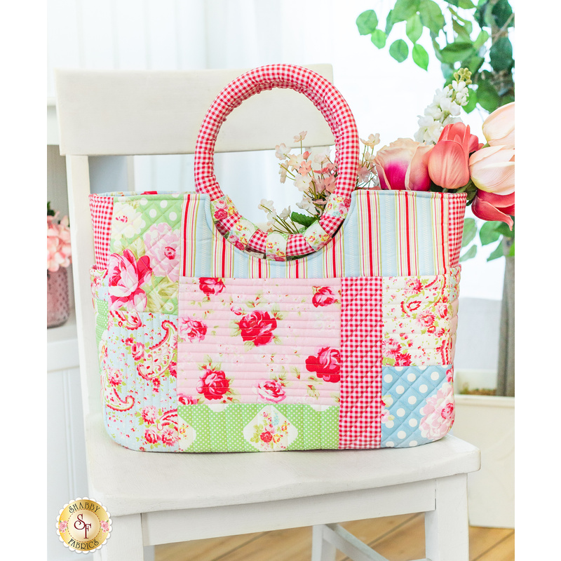 Finished tote bag made with pink, blue, and green floral fabrics filled with pink and white tulip flowers on a white chair in front of a white wall and houseplant in the background