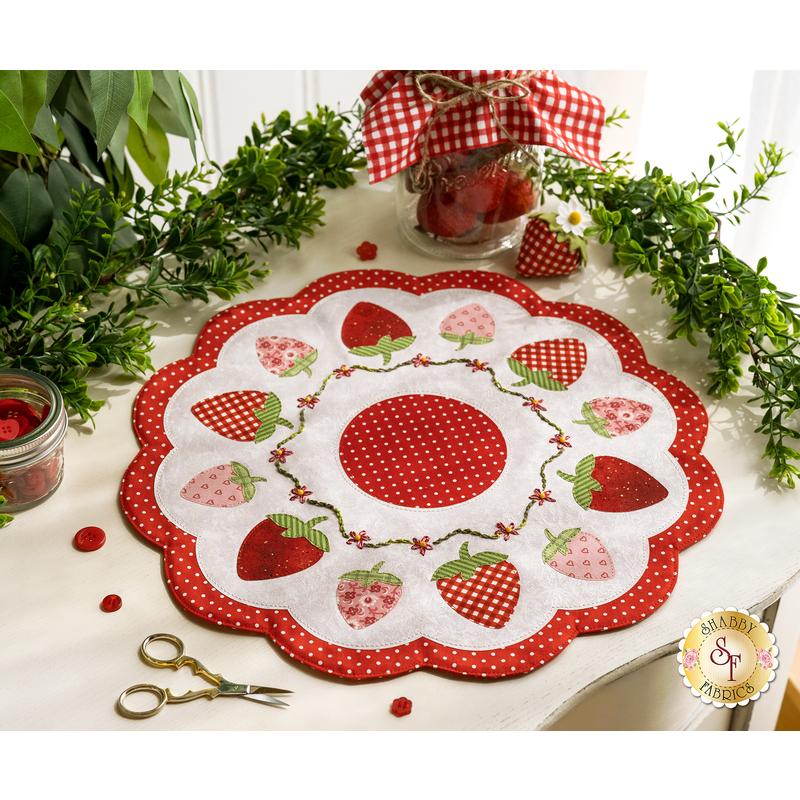 A white countertop with foliage and a houseplant in the background with a white and red strawberry themed table topper with notions and a jar of strawberries in the background