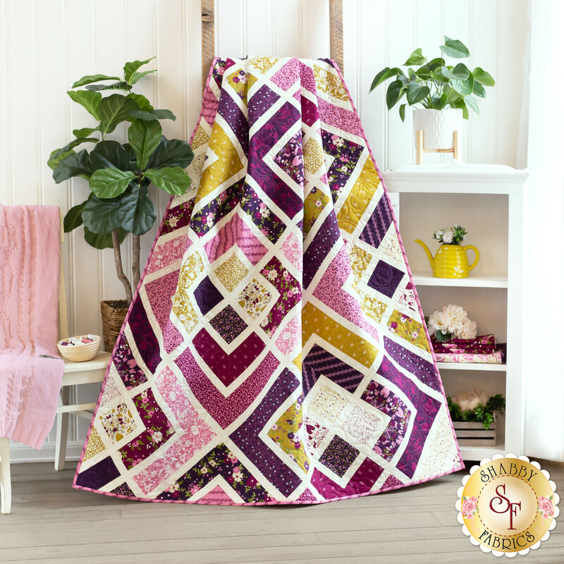 Image of geometric quilt gathered and draped in front of a white cabinet with a houseplant and yellow teapot  and a white chair with a pink blanket draped over it and a green houseplant in the background.