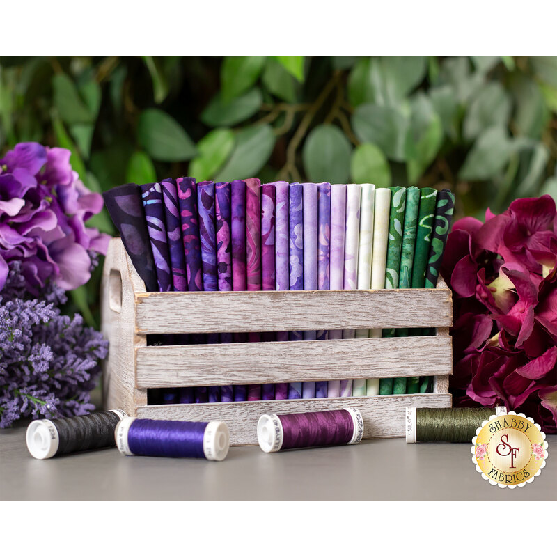 black, purple, fuschia, and green floral batik fabrics in a wood basket, surrounded by flowers and greenery