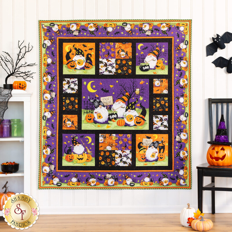 A Halloween-themed quilt hanging on a white paneled wall with Halloween decor on a white shelf and small black chair