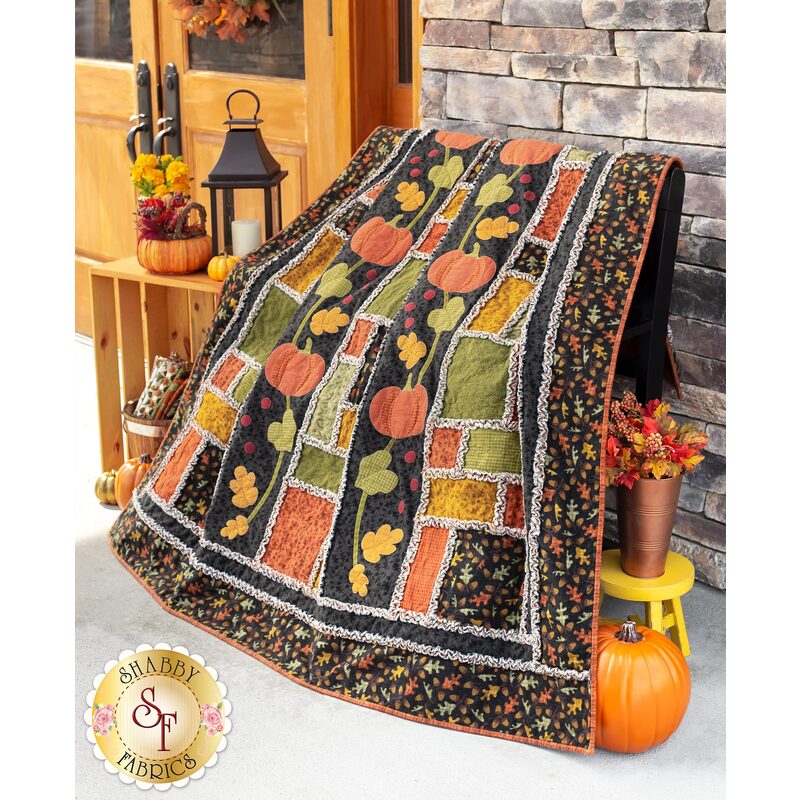 Large autumn themed quilt draped over furniture with a pumpkin and small stool with flowers on the floor in front of a small wooden shelf with a lantern and decor on it in front of wooden double doors.
