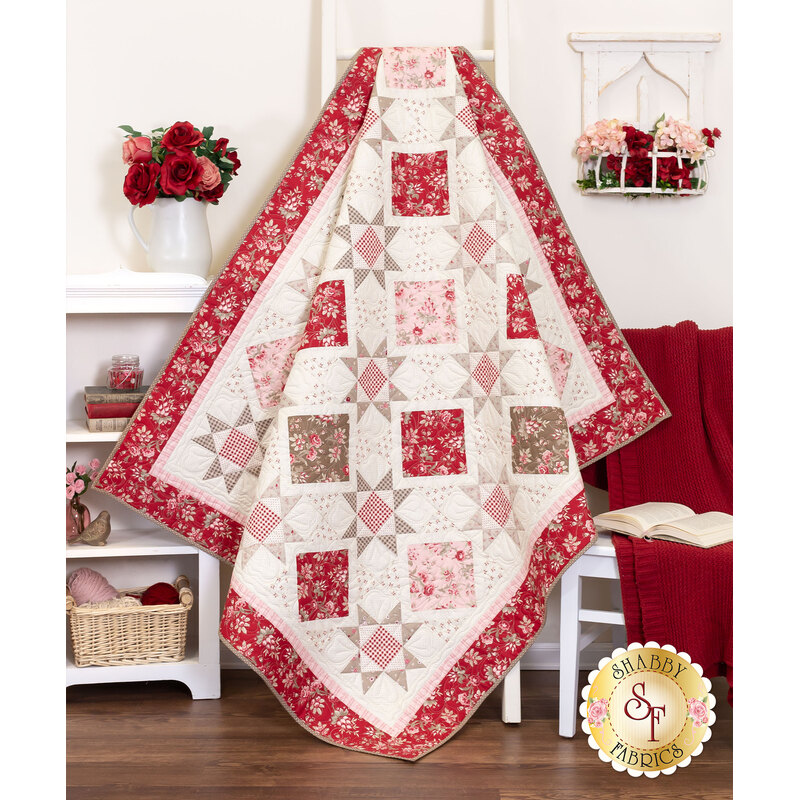 Photo of a red, pink, and cream quilt made with traditional style quilt star blocks and diamond blocks draped against a white paneled wall with white furniture and red and pink decor like a blanket, books, roses, and yarn.