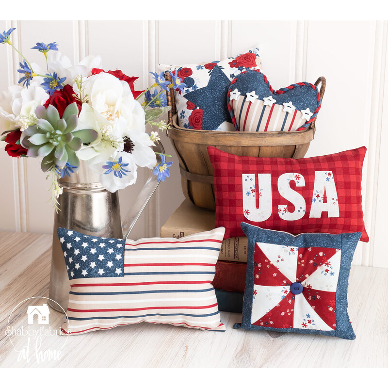 A white countertop and white paneled wall with a collection of small patriotic themed pillows in the foreground with a basket full of more pillows and a metal pitcher filled with flowers to the left.