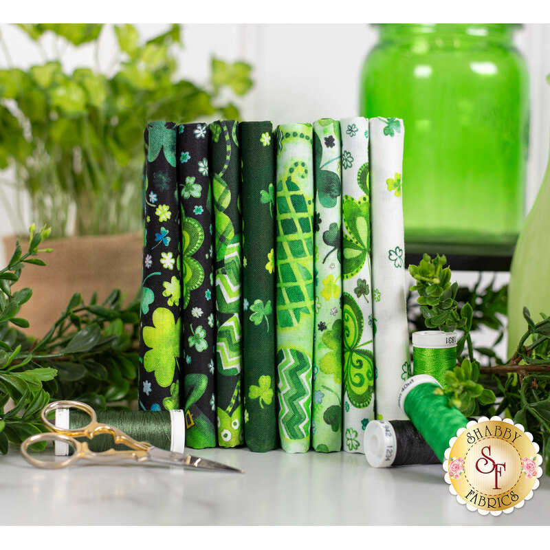 Green shamrock themed fabrics stacked on a table with green thread and plants