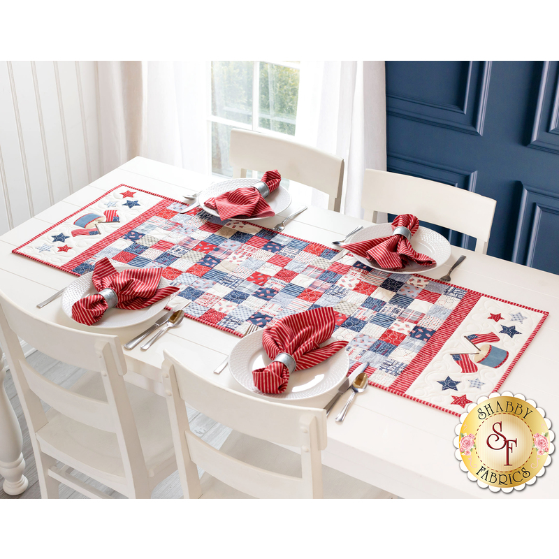 Photo of a patriotic red white and blue table runner in a patchwork pattern on a white table with four place settings, each with matching red napkins in rings, and a bright window in a blue wall in the background
