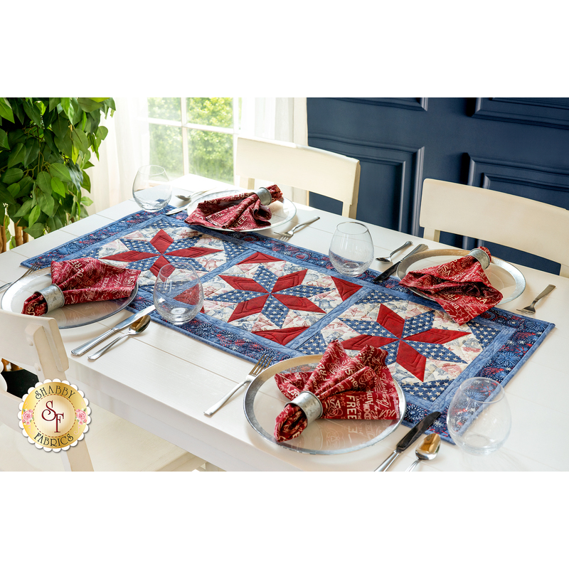 Red, white and blue table runner in the center of a white table with four chairs and place settings with matching fabric napkins in rings and a plant, window, and dark blue wall in the background