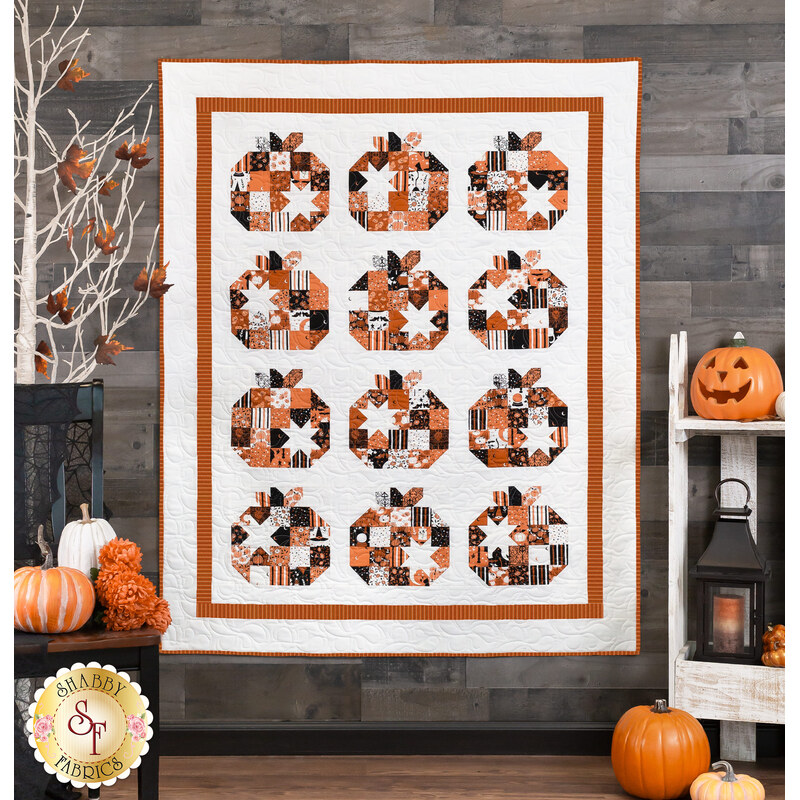 A photo of a pumpkin and Halloween themed quilt hanging on a gray paneled wall with halloween decor all around