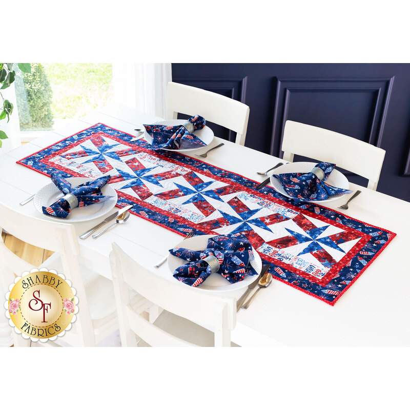 Patriotic themed red white and blue table runner on a white table with four place settings with matching cloth napkins all around with four white chairs and a bright window and blue wall in the background