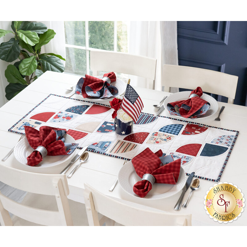 A white table in front of a blue wall with a bright window and green houseplant in the background with four place settings and a patriotic themed table runner with matching cloth napkins at each place setting and an American flag centerpiece.