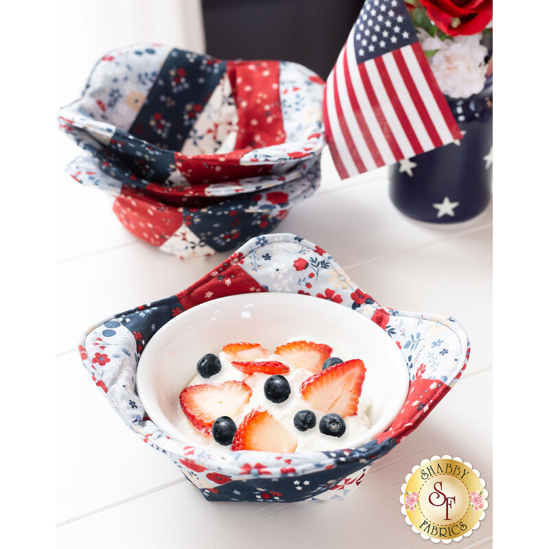 A red, white, and blue bowl cozy made with patriotic fabrics holding a white bowl filled with fruit and whipped cream on a white table with a stack of matching bowl cozies in the background with a patriotic centerpiece on the right.