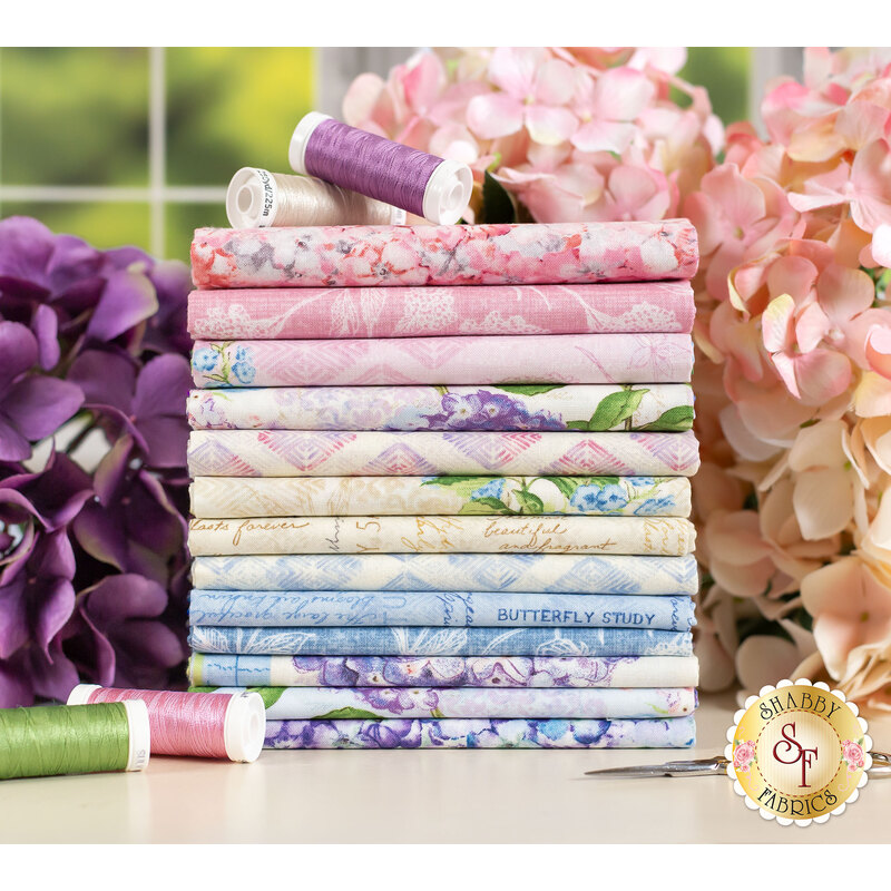 pink, blue, and cream floral fabrics in the Hydrangea Mist FQ set stacked on a table and surrounded by hydrangeas