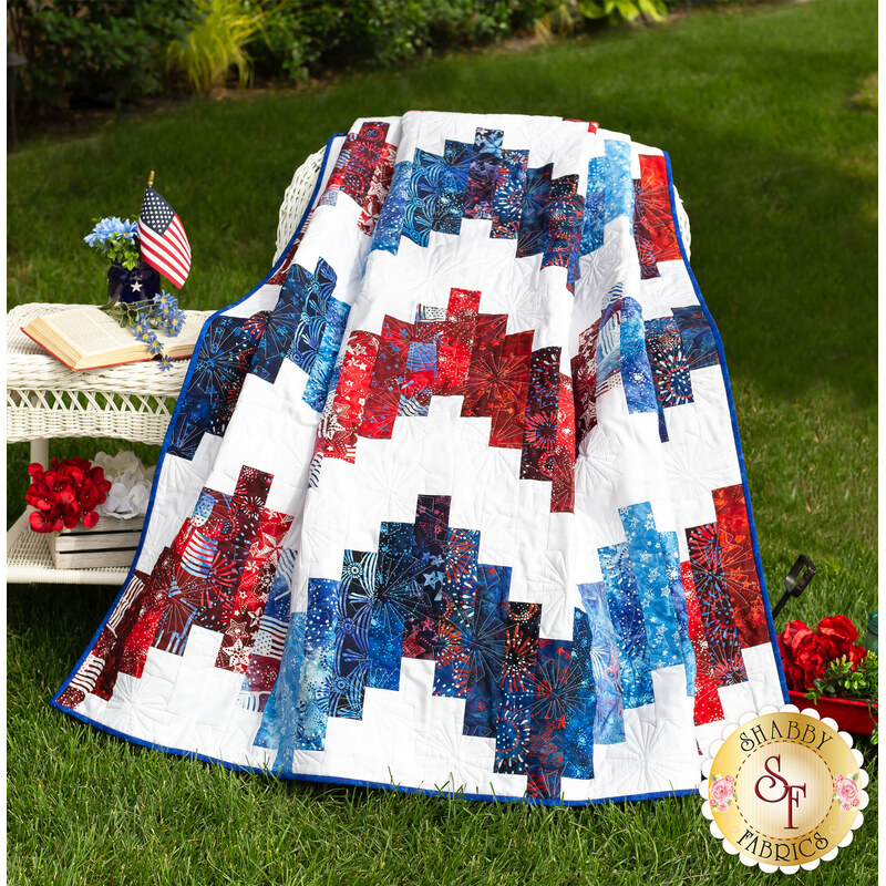 Gathered red white and blue quilt next to a white wicker table with red flowers and a book and green grass all around