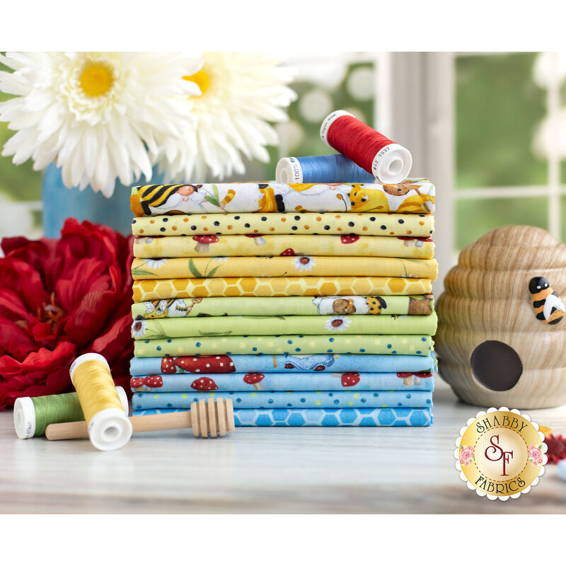 Yellow, green, and blue fabrics featuring gnomes, bees, flowers, red mushrooms, and honeycomb prints stacked on a table with flowers and a beehive