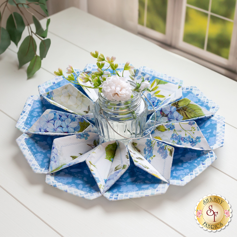 Flower shaped table topper with central opening containing a glass jar filled with fresh flowers. Topper is made of white and blue floral fabrics with green accents.