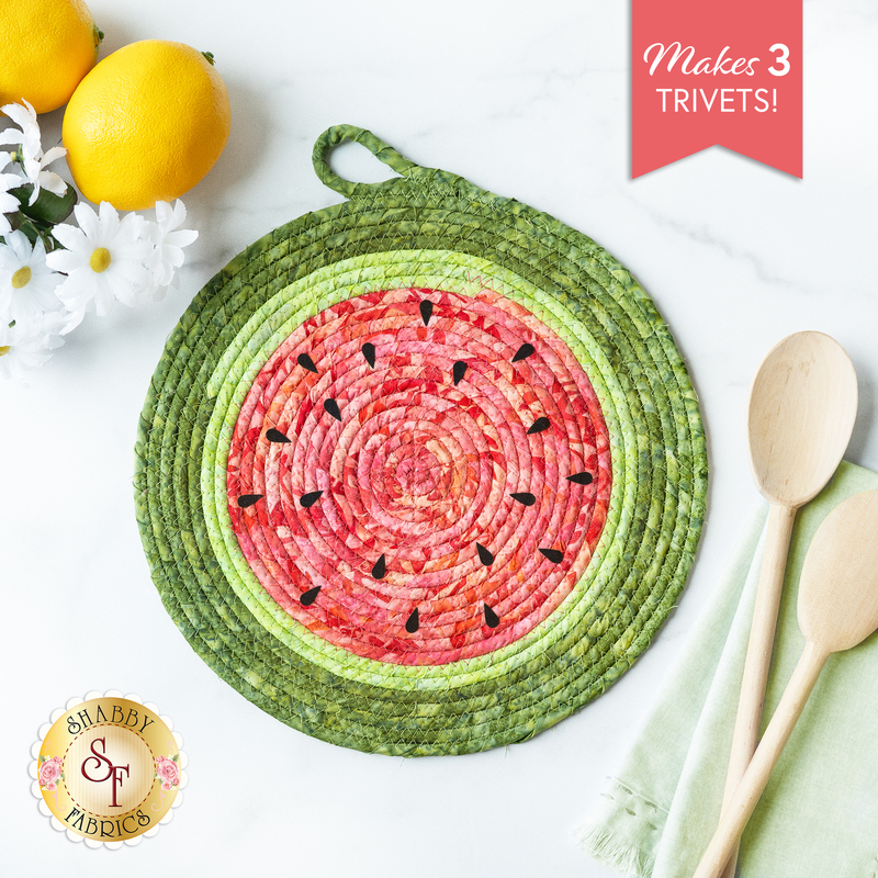 Round Watermelon Trivet made with pink and green batiks on a white countertop with lemons, daisies, and wooden spoons on either side