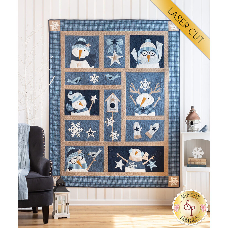 A blue and tan quilt featuring several snowmen hanging on a white panel with a small white shelf with winter decor and a black armchair to the left with a white vintage lantern on the floor