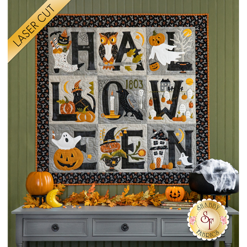 Photo of a small Halloween quilt hanging on a green paneled wall with fall/Halloween decor and a gray table with drawers. Each block of the quilt contains a letter of the word 