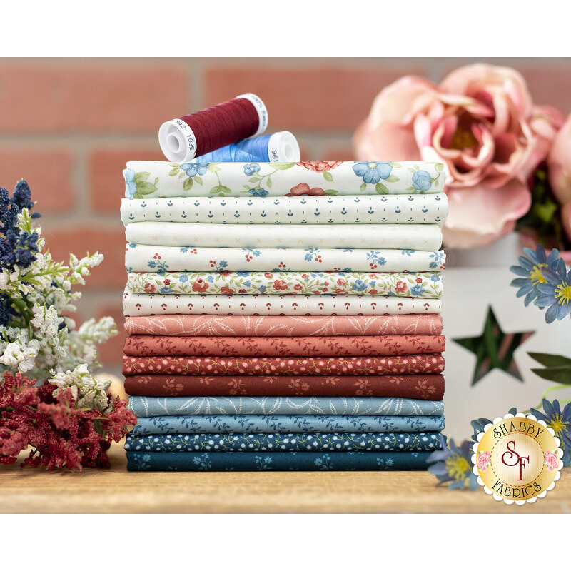 Muted red and blue fabrics and creamy whites, printed with florals, on a wood table in front of a red brick wall surrounded by flowers
