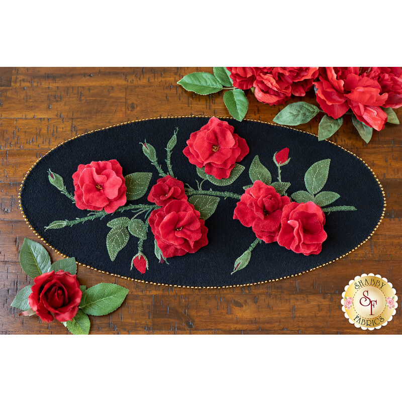 A black oval table topper with 3-dimensional roses and green leaves with gold beads around the edge on a brown wooden table with red roses on the top and bottom