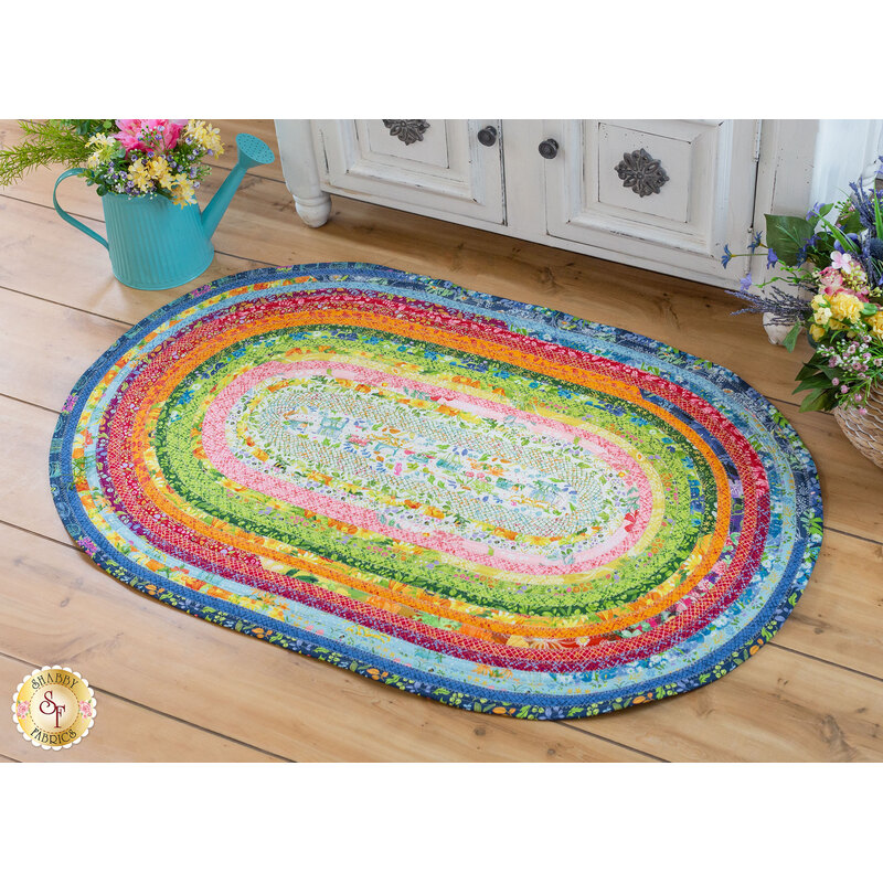 Colorful jelly roll rug with blue, pink, orange, yellow, green and white concentric circles on a wooden floor with a white cabinet and teal watering can full of flowers in the background