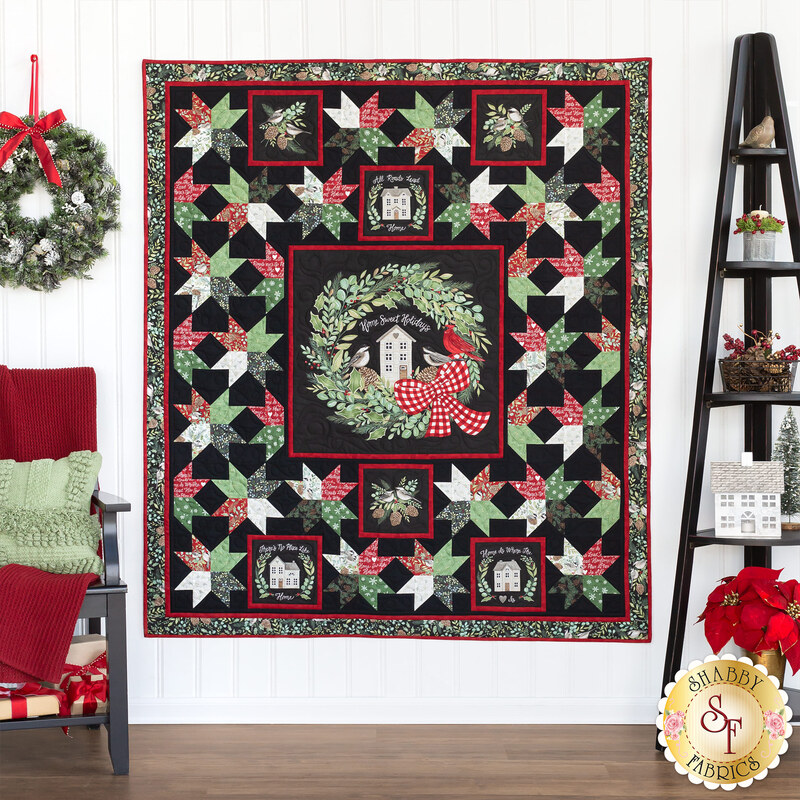 A finished Christmas Eve Quilt hanging on a white paneled wall with a ladder shelf on the right featuring homey decor, and a chair with a red blanket and green pillow on the other with a wreath hanging on the wall.