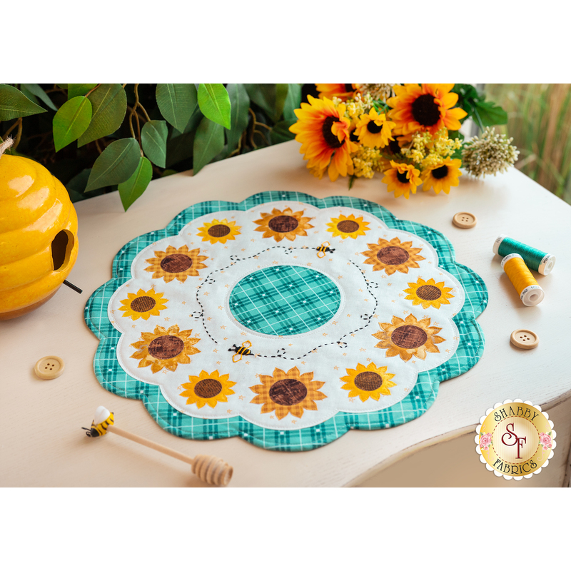 A white scalloped table topper with aqua plaid edges and center with simple applique sunflowers in a ring, accented by hand embroidered bee details on a white tabletop with honey and bee themed decorations, thread spools, buttons, and sunflowers with a green plant in the background
