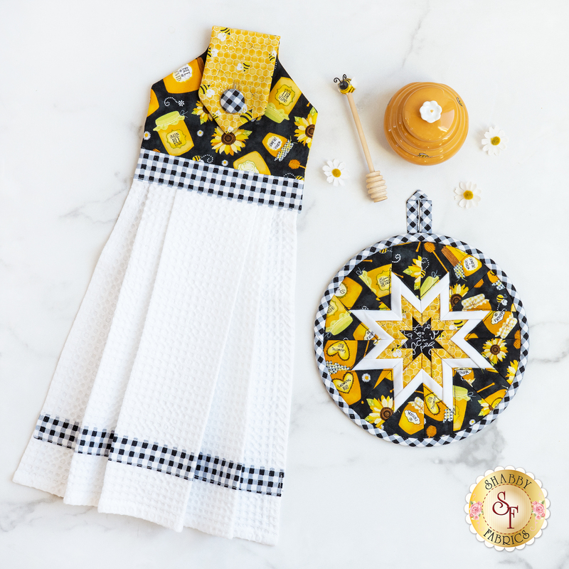 Hanging towel and hot pad for August, featuring jars of honey, sunflowers, and gingham with bee themed decorations scattered around.