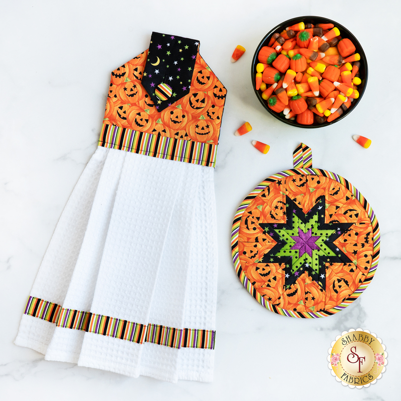 Hanging towel and hot pad for October, jack-o-lanterns, and stars with striped accents on a white countertop with a bowl of candy corn in one corner
