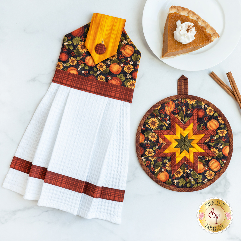 Hanging towel and hot pad for November, featuring pumpkins, sunflowers, and bright yellow textures with plaid accents on a white countertop with a plate with a slice of pumpkin pie on it