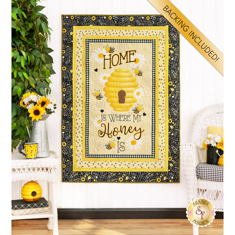 Photo of finished panel quilt hanging on a white paneled wall with a houseplant on one side, sunflower and bee decor, and white wicker furniture with a banner in the top right corner that reads 
