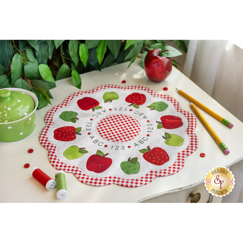 Scalloped table topper on a white counter featuring a ring of red and green apples on a white background with the alphabet and numbers in an embroidered ring in the middle. Pencils, spools of thread, a pot, buttons, and greenery are on the countertop 