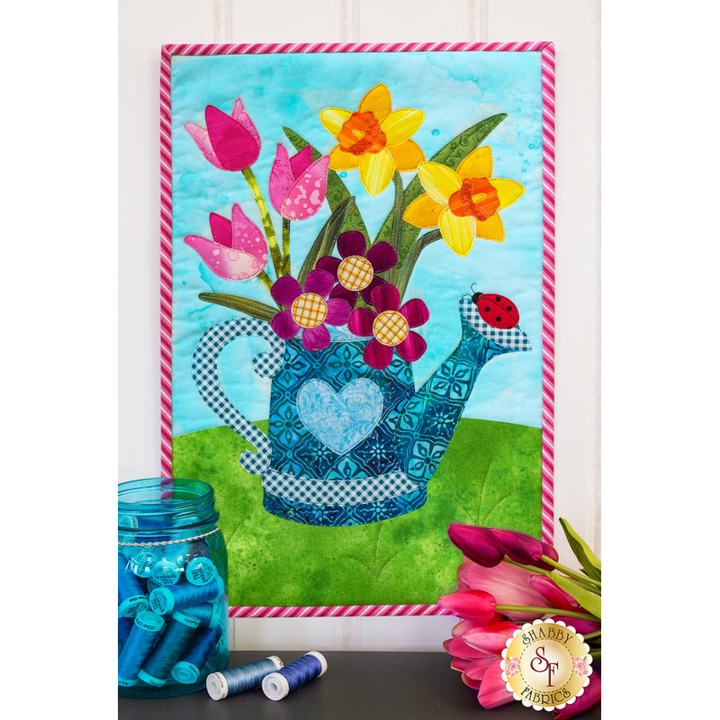 A photo of a bright, spring-themed wall hanging featuring a watering can filled with flowers and a jar of thread with pink tulips on the countertop in the foreground.