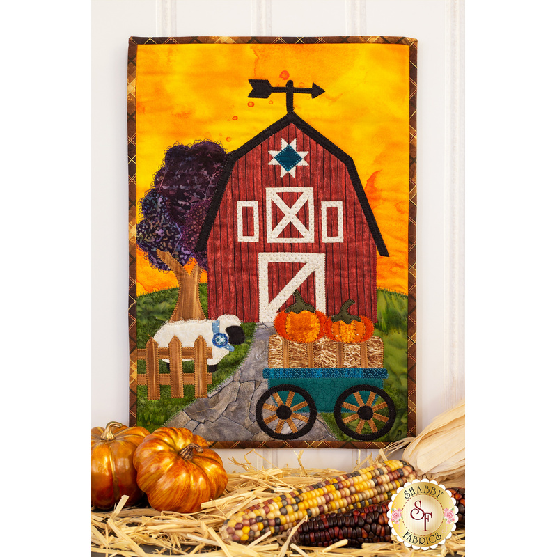 A photo of an autumn-themed wall hanging featuring a red barn, wagon full of pumpkins, and a sheep hanging on a white wall with fall decor of straw, dried corn, and small pumpkins in the foreground.