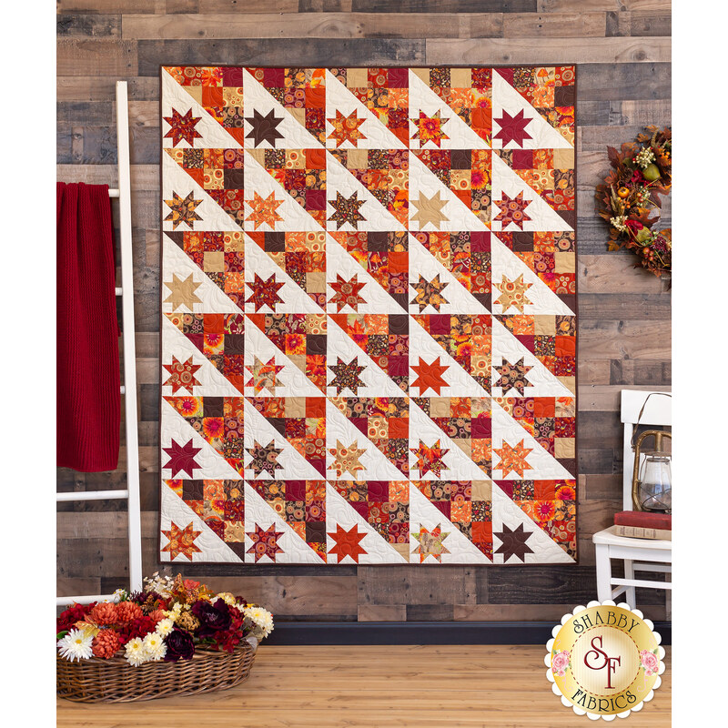 Photo of a geometric quilt with small stars in each block, made with autumn themed fabrics hanging on a dark wood paneled wall with autumn floral decor and painted white furniture on either side.