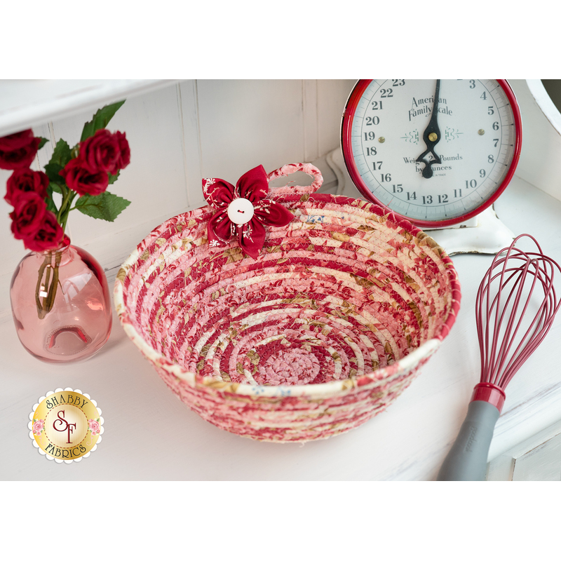 Photo of a bowl made from coiled red and cream colored fabrics with a red fabric flower sitting on a white countertop with a red whisk, scale, and pink bottle with red roses in it.