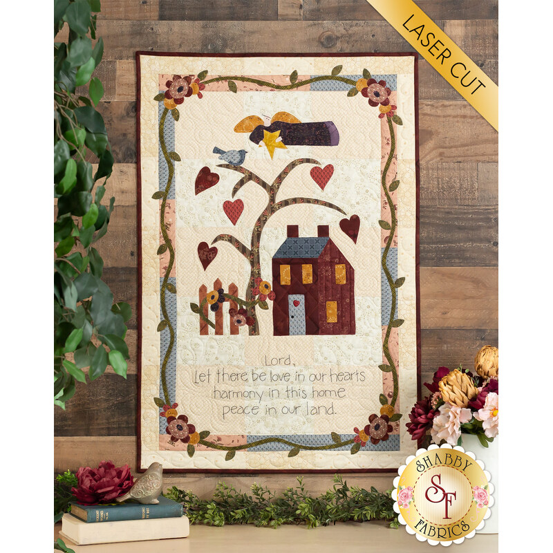 Photo of a small quilt hanging on a brown paneled wall featuring a house, tree bearing hearts, an angel flying overhead, and the words 
