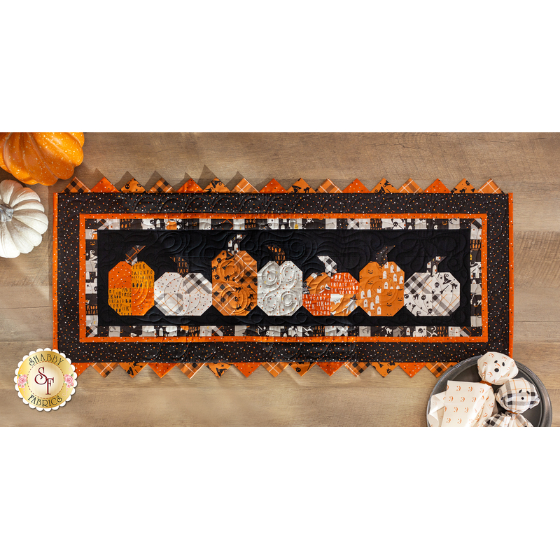 Photo of a Halloween themed table runner made with black, orange, and white fabrics with points along the top and bottom and a row of pumpkins in the center, on a wooden table top with pumpkins and Halloween decor