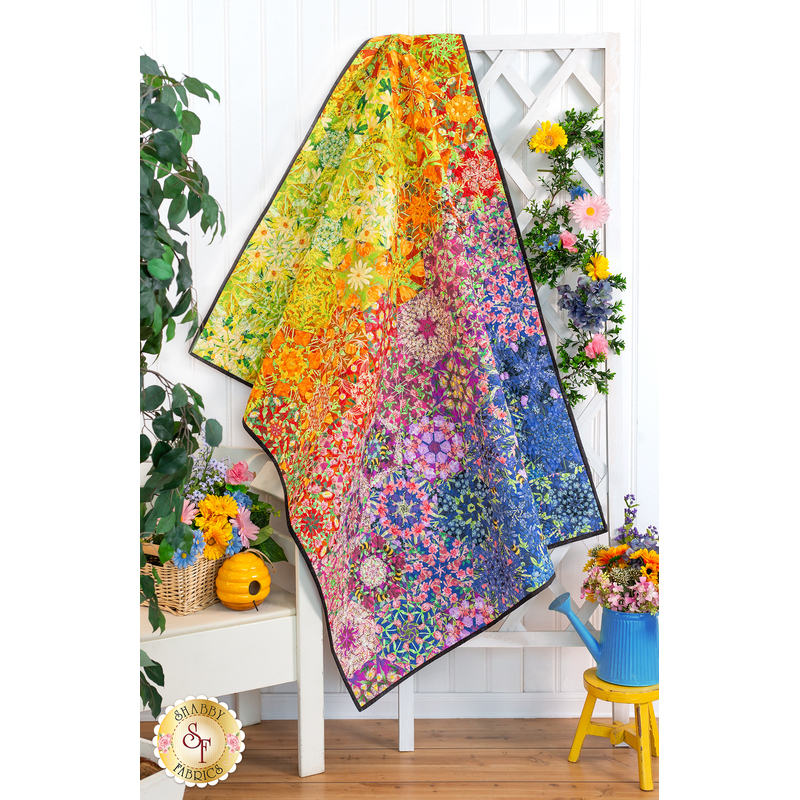 Photo of a rainbow colored floral quilt draped from a white lattice with a white bench and floral decor all over