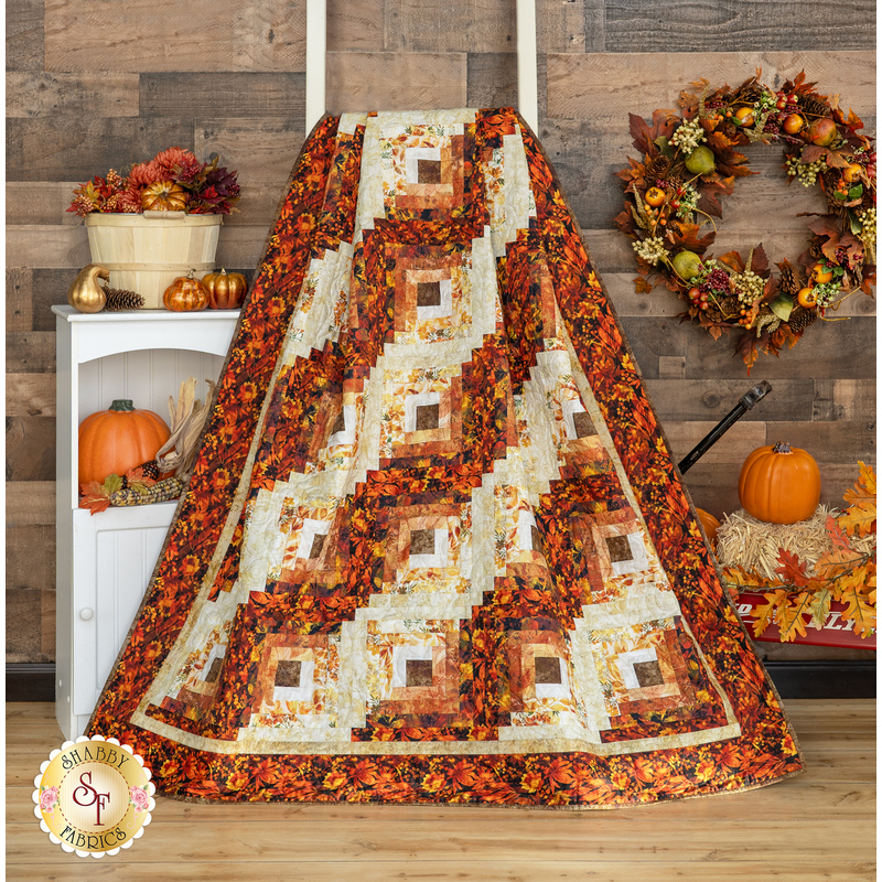 Photo of the Reflections of Autumn Log Cabin Throw Quilt draped over a white ladder in front of a dark paneled wall with a small white shelf to one side, an autumn wreath, and pumpkin decor