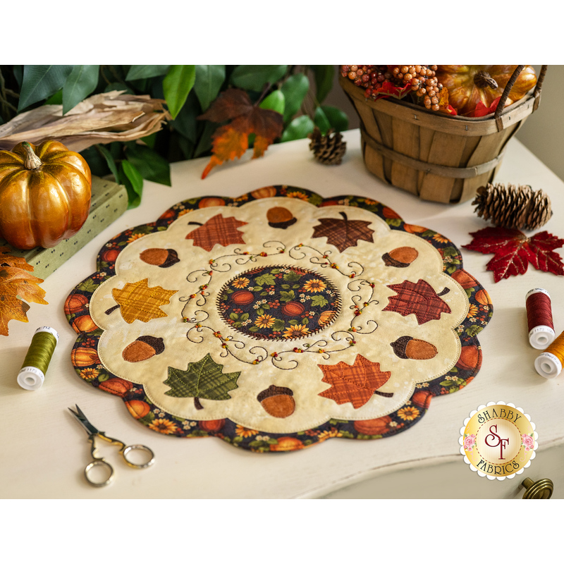 Photo of an autumn themed table topper on a white table with fall decor all around including leaves, a gold pumpkin, and sewing notions