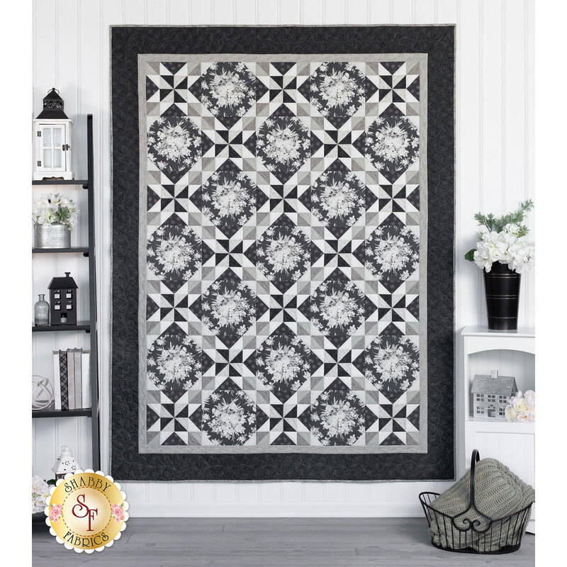 Photo of a black, white, and gray geometric quilt hanging flat on a white wall in a room with monochromatic decor such as a basket, gray blanket, and white flowers in a tall black vase.