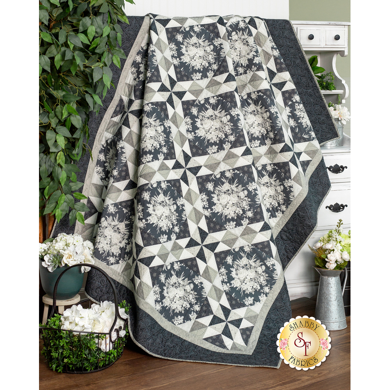 The completed Lattice Bouquet Quilt in Steelworks, rich shades of cool, smoky, and medium gray fabrics. The quilt is artfully draped from an armoire and staged with coordinating decor and leafy green foliage.
