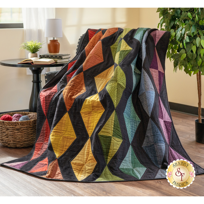 Photo of the Scintillation flannel quilt made with rainbow colored fabrics and black accents draped over a chair in a brightly lit room with a basket of yarn and a table with a lamp, plant, and books in the background