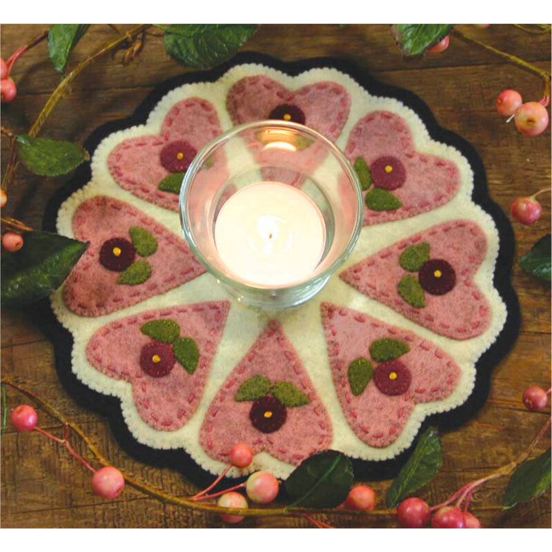 Photo of a small scalloped circular mat on a brown table with berries and vines all around it. On the mat is featured large stylized pink hearts and red flowers with a lit candle at the center.