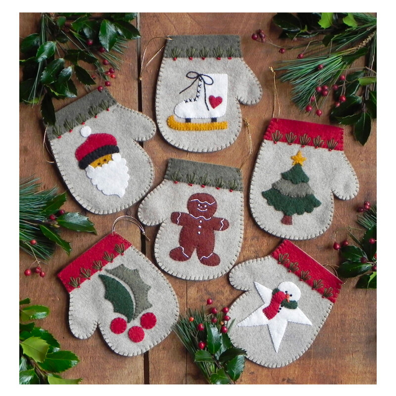 Photo of 6 mini mitten ornaments on a brown table top with pine tree boughs. Each mitten has a different motif including holly, gingerbread, and an ice skate
