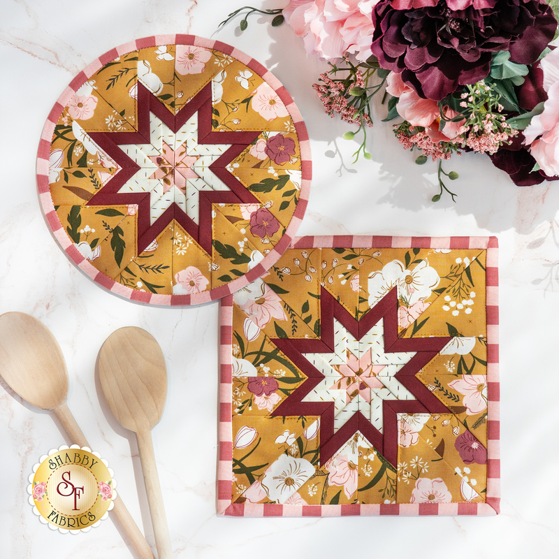 Photo of two folded star hot pads, one round and one square, made with yellow, white, and red floral fabrics laying flat on a white countertop with wooden spoons and a bouquet of flowers